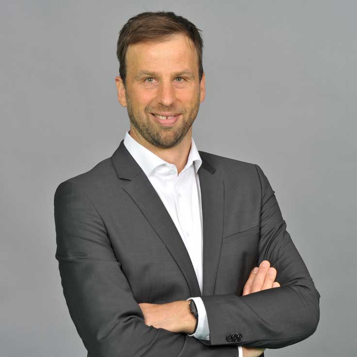 A man in a grey suit stands with arms crossed against a grey background. Portrait of Stefan Nass.