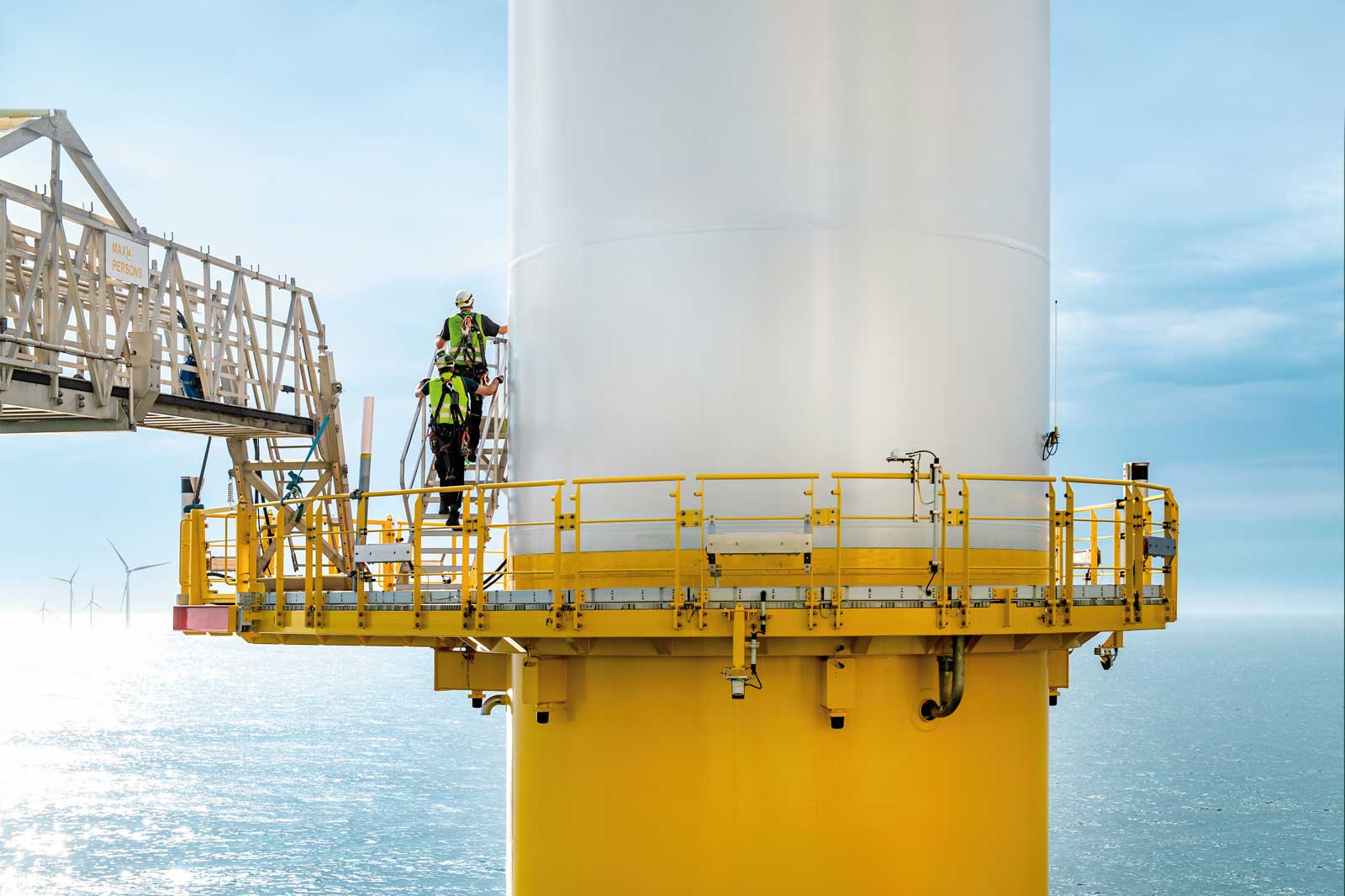 A worker is climbing an offshore wind turbine, surrounded by calm waters and a clear sky.