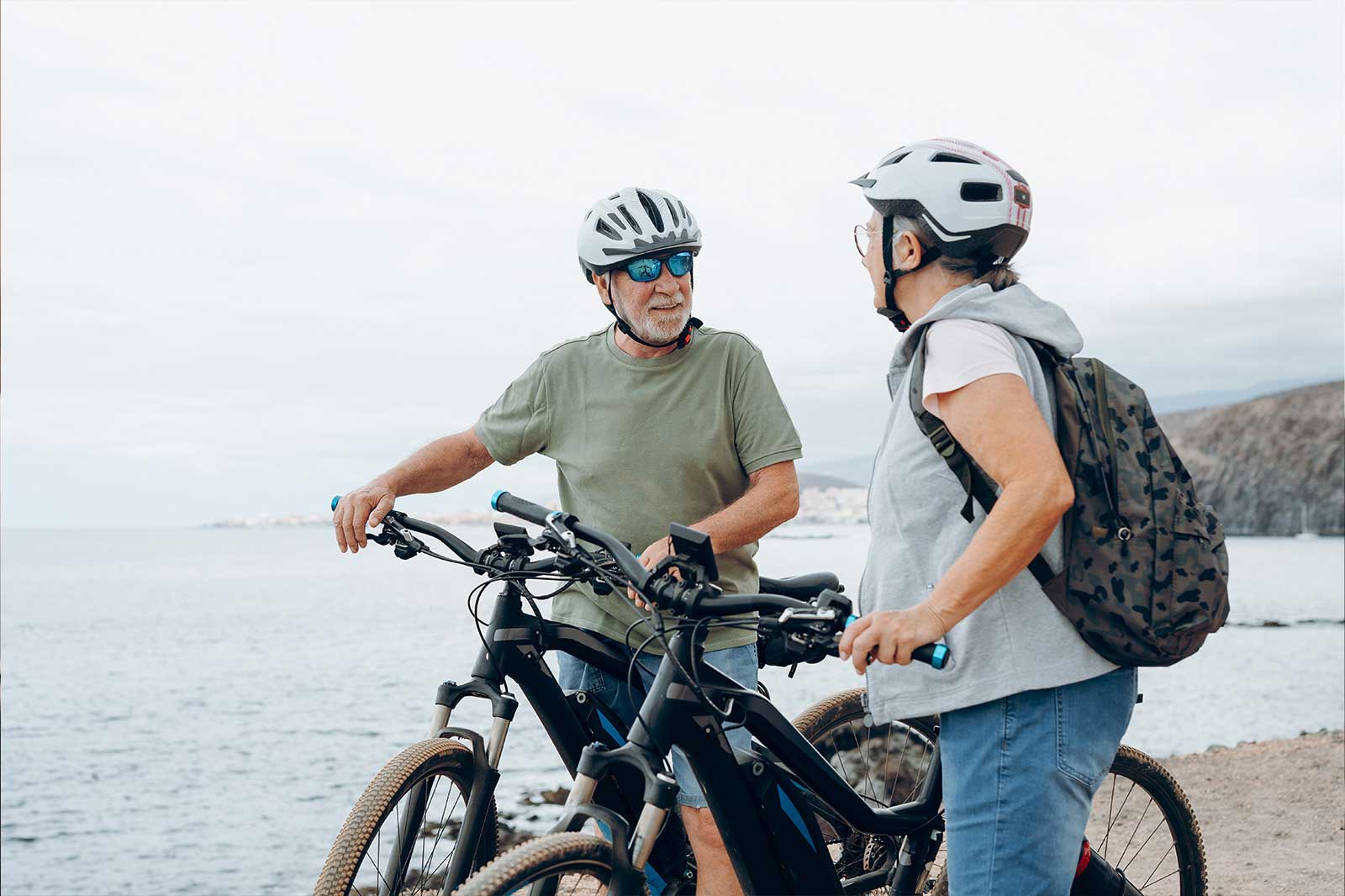 Ein älteres Paar steht an einem Gewässer, jeder mit einem E-Bike, und unterhält sich. Im Hintergrund sind sanfte Hügel.