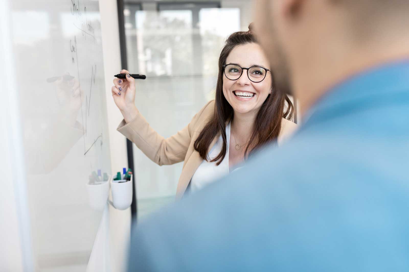 Eine Frau präsentiert an einem Whiteboard, während ein Mann im Vordergrund zuhört. Sie erklärt Diagramme und Notizen.
