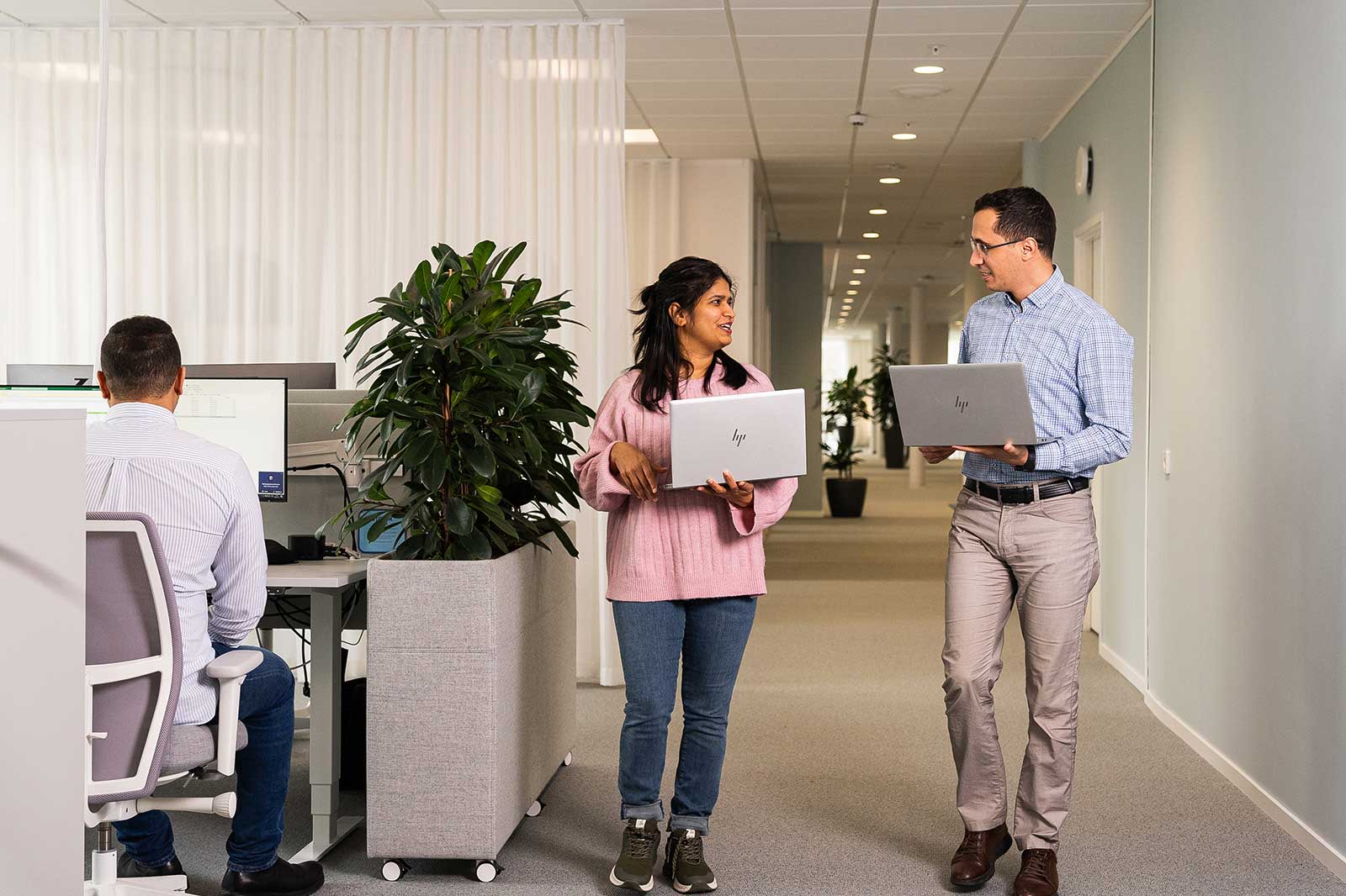 Zwei Personen in einem modernen Büro gehen mit Laptops und unterhalten sich. Im Hintergrund sind Pflanzen und Arbeitsplätze sichtbar.