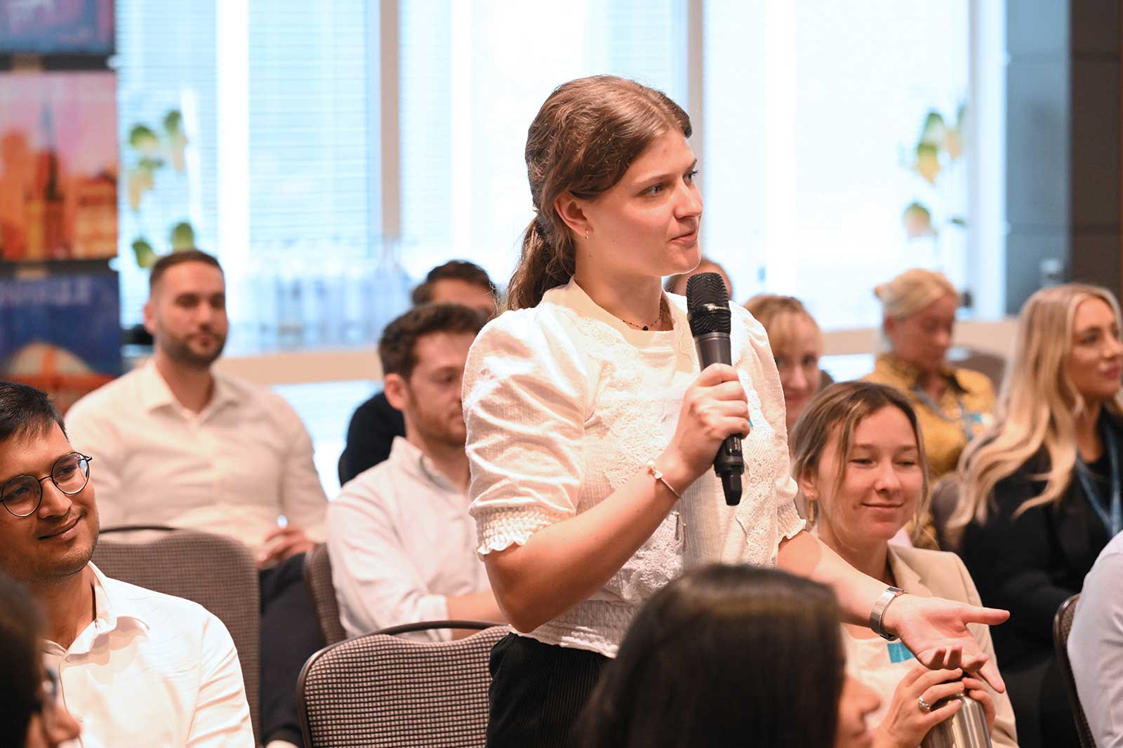 Eine Frau stellt eine Frage auf einer Konferenz und hält ein Mikrofon, während im Hintergrund sitzende Zuhörer zu sehen sind.
