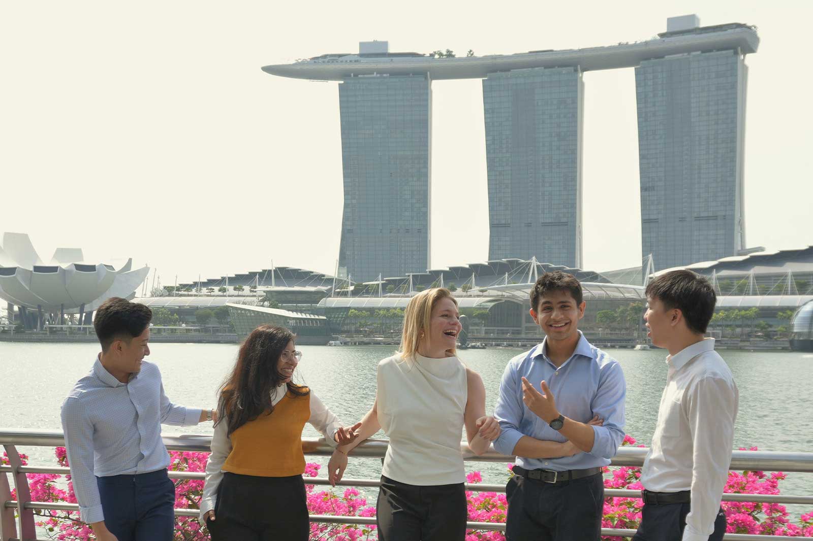 Eine Gruppe von fünf Personen steht am Wasser mit dem Marina Bay Sands im Hintergrund und rosa Blumen im Vordergrund.