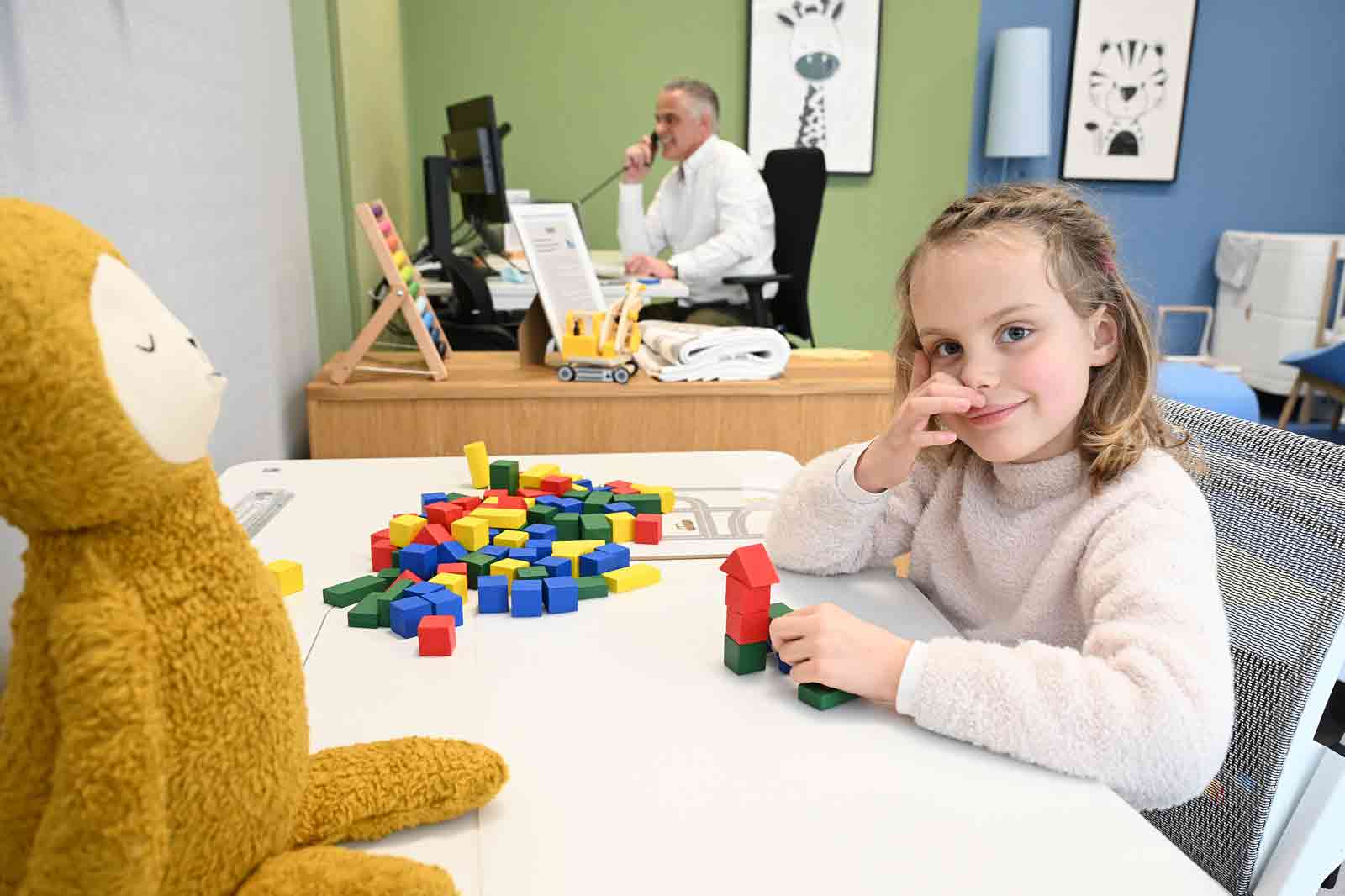 Ein Kind sitzt an einem Tisch mit bunten Bauklötzen, während ein Plüschaffe daneben steht. Im Hintergrund arbeitet ein Erwachsener.