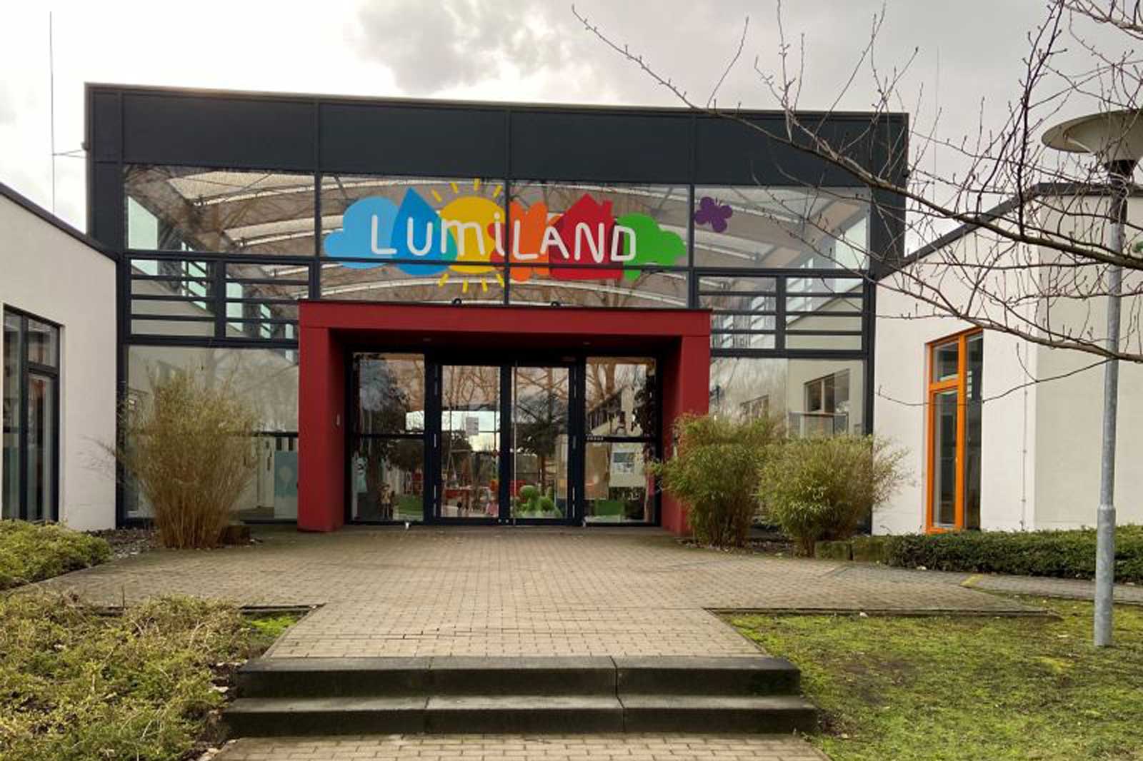 Das Gebäude von Lumiland mit buntem Logo über dem Eingang und Pflanzen im Vordergrund. Die Fenster sind groß und modern.