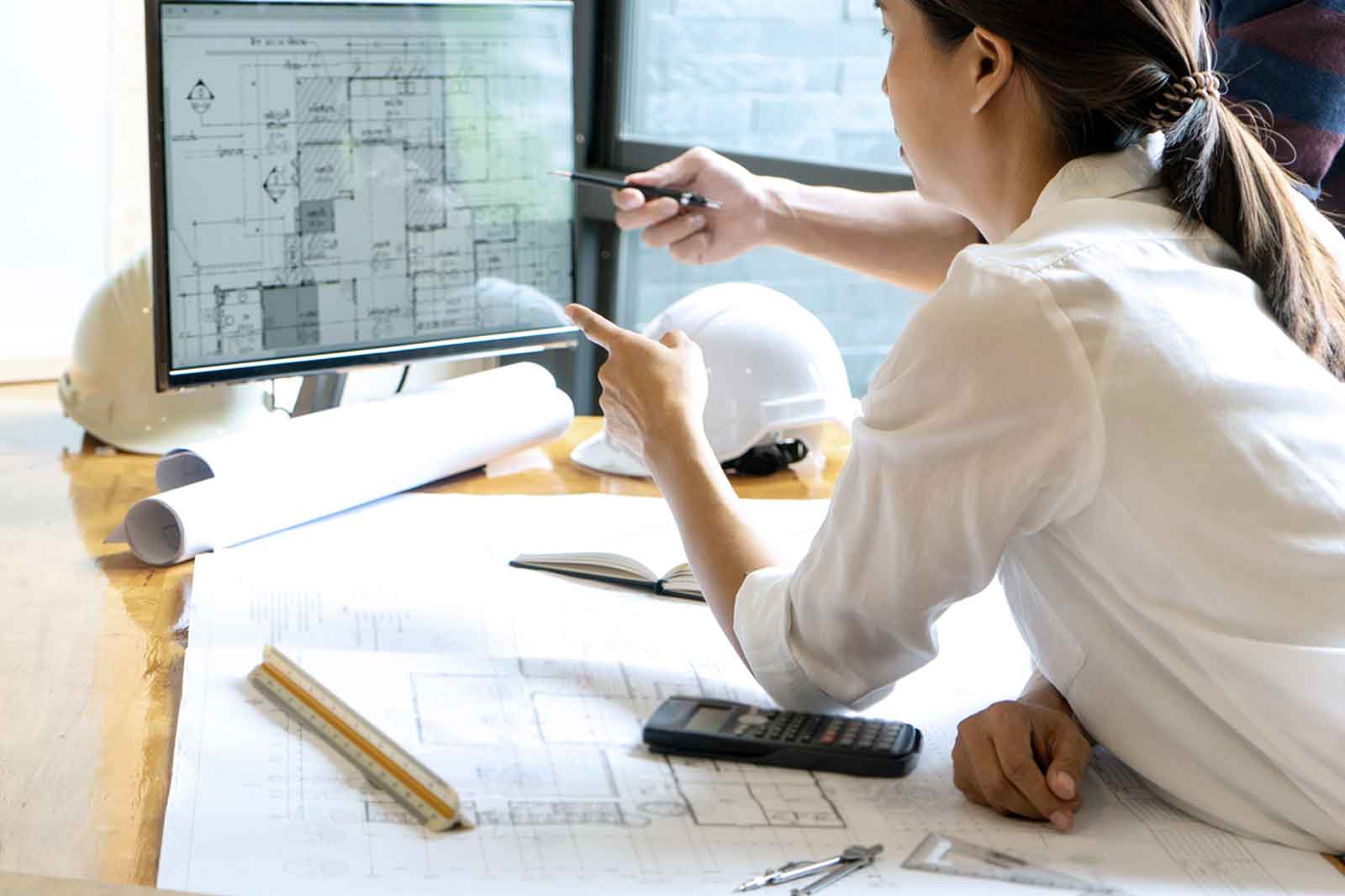 Eine Person überprüft architektonische Pläne auf einem Computerbildschirm und zeigt auf die Blaupausen auf dem Tisch.