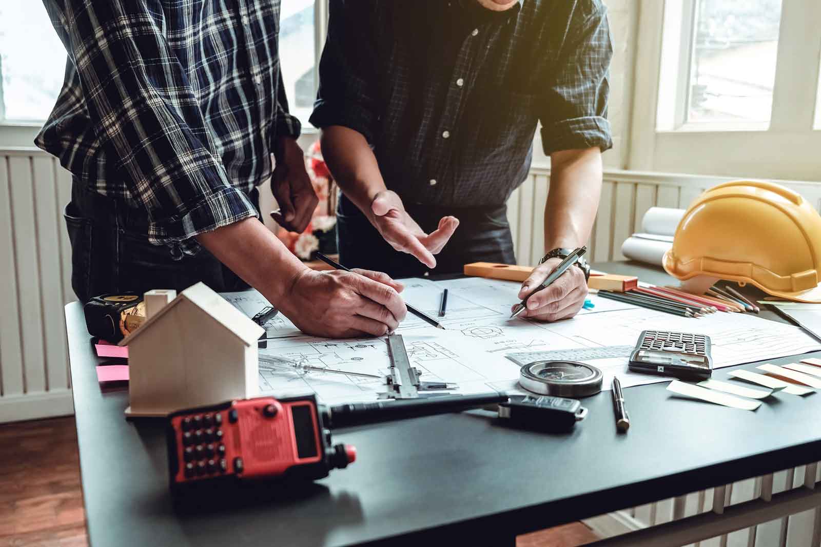 Zwei Personen arbeiten über Bauplänen an einem Tisch, mit Werkzeugen und einem Schutzhelm in der Nähe.