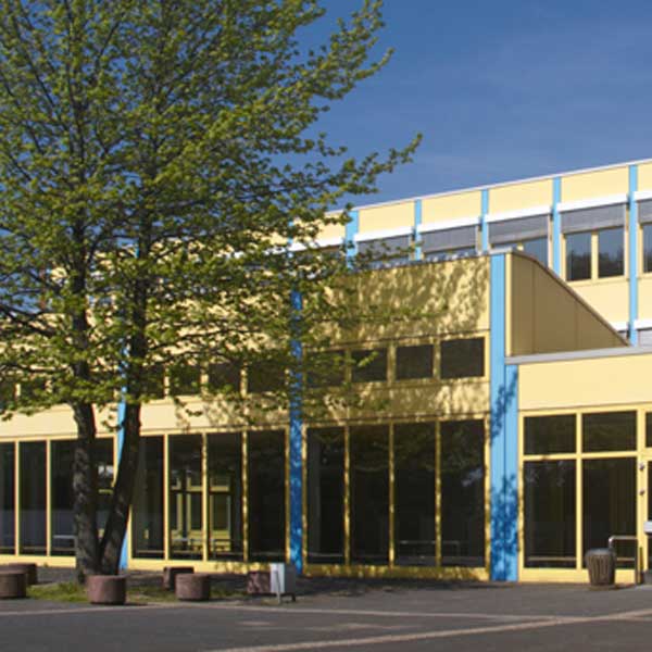 Moderne Gebäudeaußenansicht mit großen Glasfenstern, gelben und blauen Akzenten sowie einem nahegelegenen Baum unter blauem Himmel.