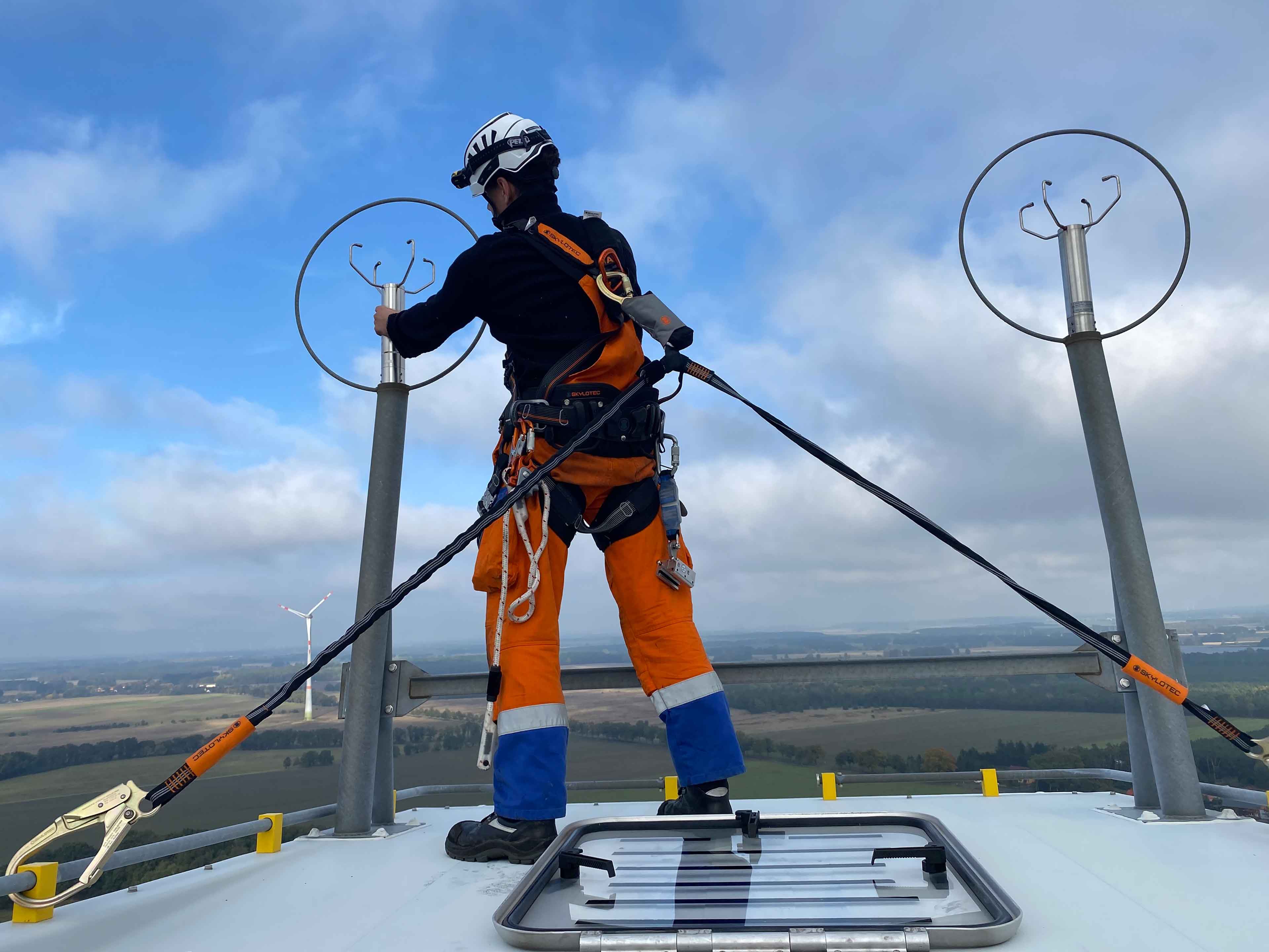 Ein Techniker in orangefarbener Uniform arbeitet an Geräten auf einem Windrad, mit einer weiten Landschaft darunter.