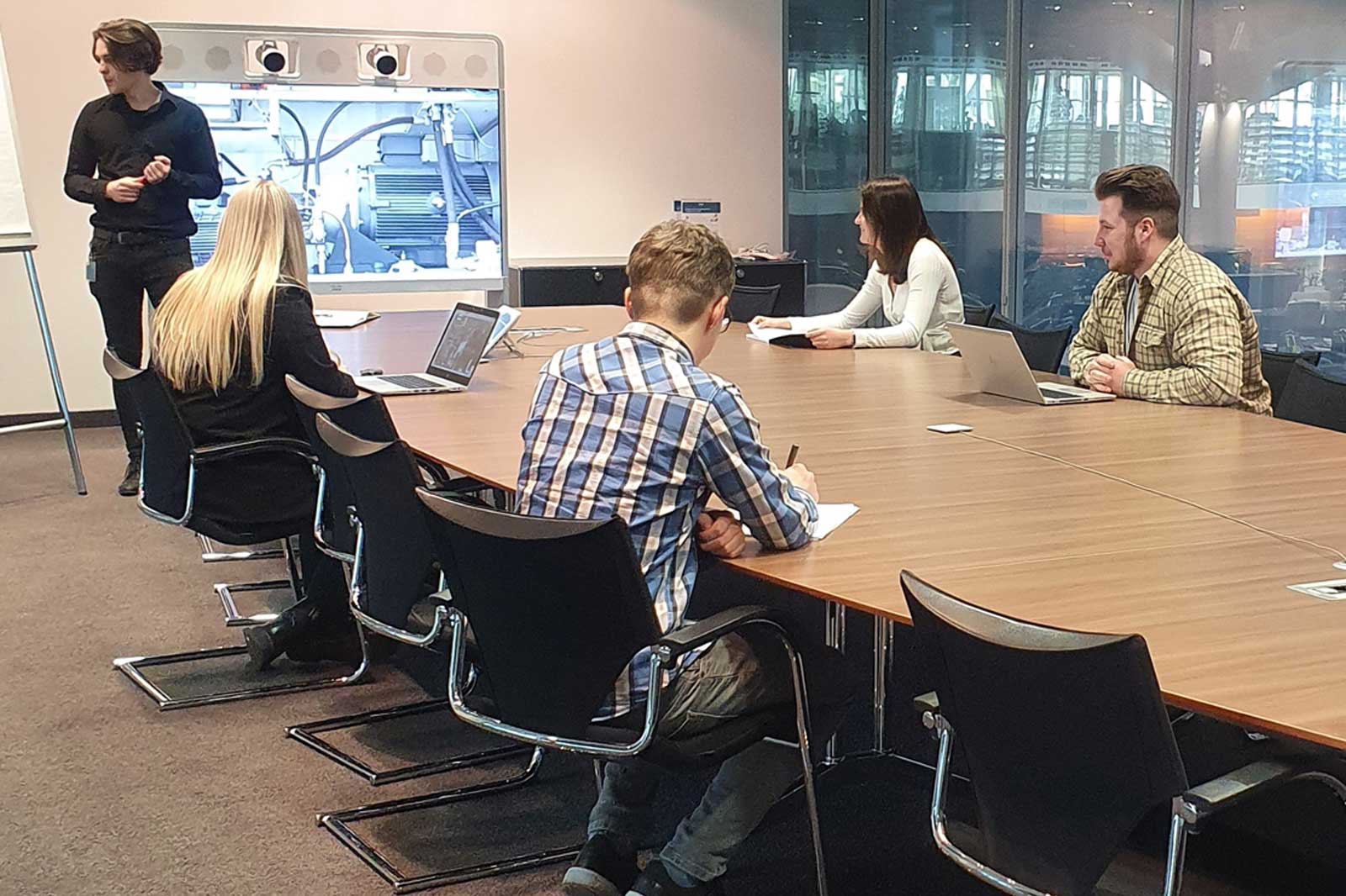 Ein Besprechungsraum mit fünf Teilnehmern, die an einem langen Tisch sitzen und auf eine Präsentation eines stehenden Sprechers achten.