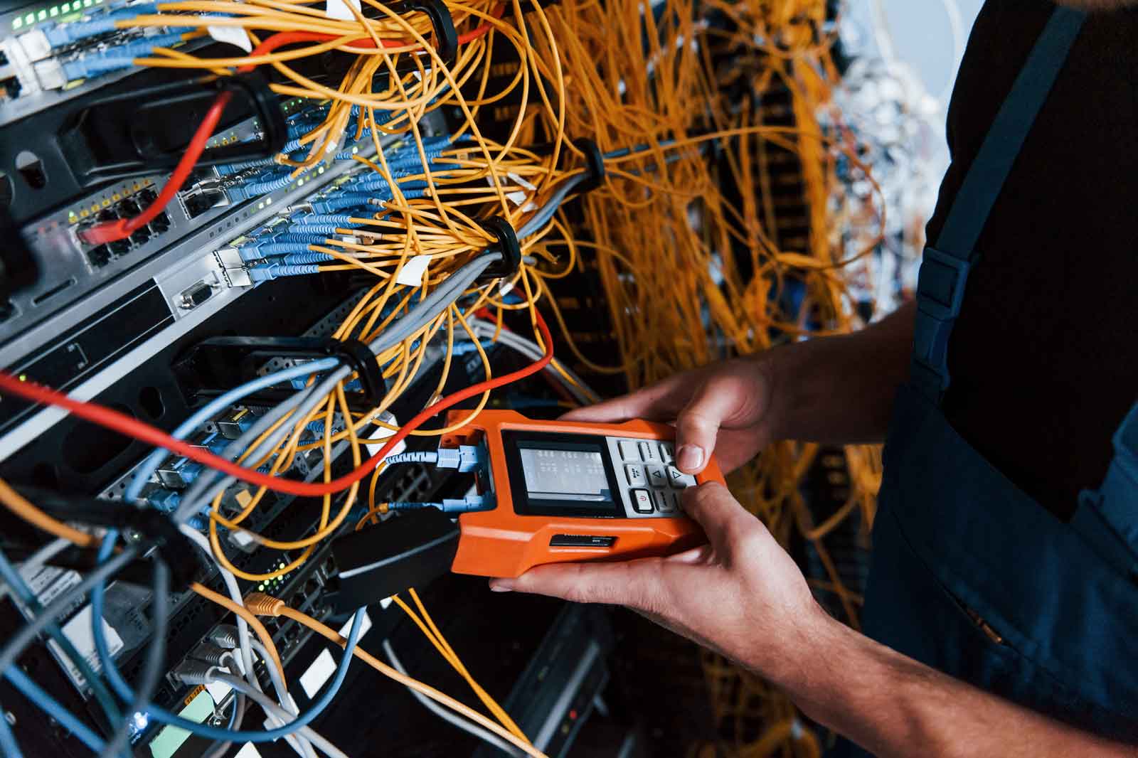 Ein Ingenieur hält ein Kabeltester inmitten eines Wirrwarrs aus gelben und blauen Netzwerkkabeln in einem Serverrack.