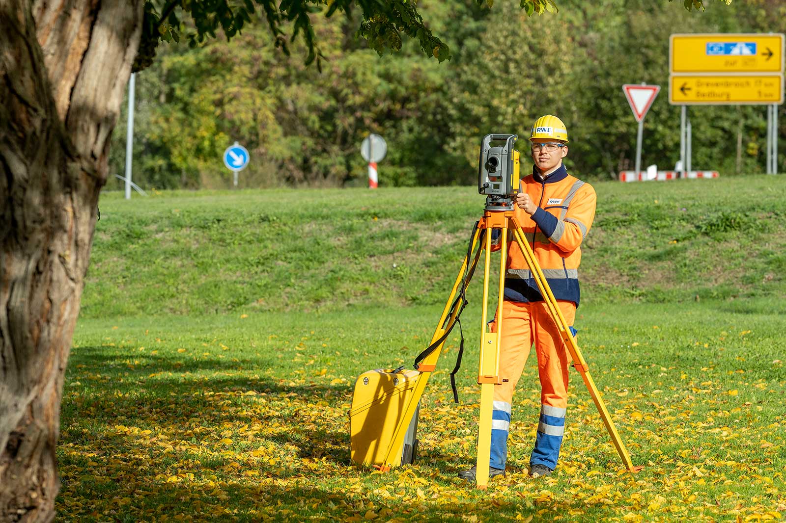 Ein Vermessungsarbeiter in orangefarbener Warnkleidung und gelbem Helm bedient ein gelbes Messgerät auf einem Stativ in einer grasbewachsenen Parkanlage mit fallenden gelben Blättern.
