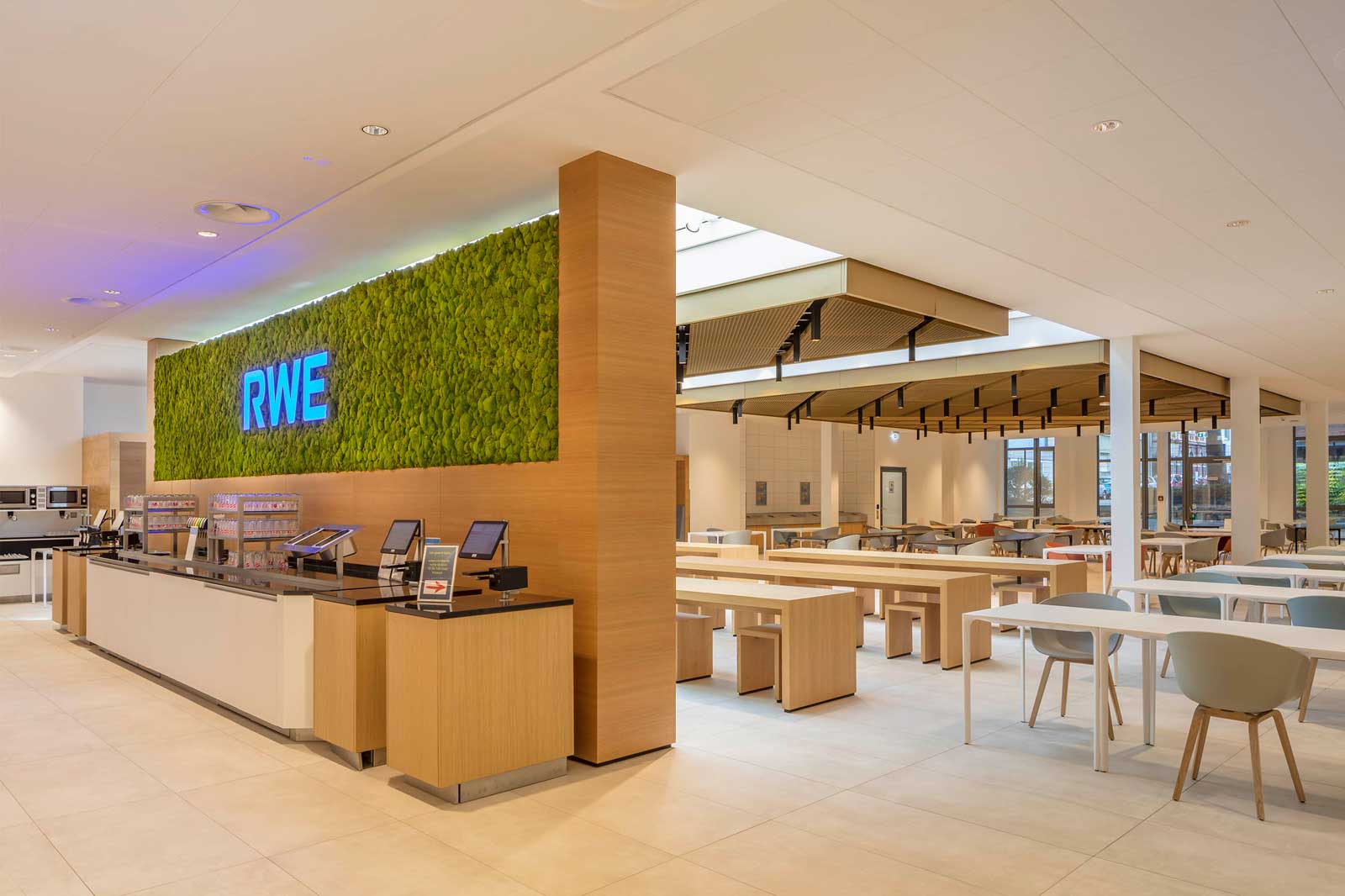 Eine moderne Cafeteria mit einer grünen Mooswand, Sitzbereichen und einem Tresen mit Selbstbedienungskiosken.