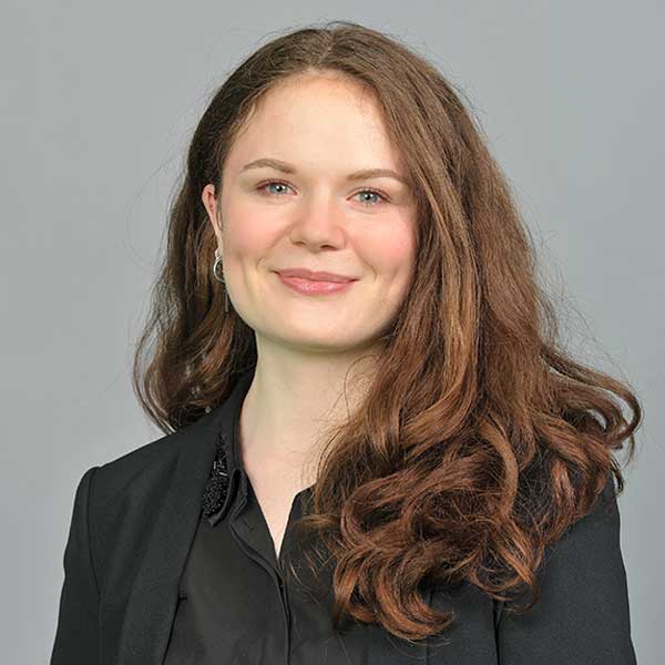 Eine Person mit lockigem, kastanienbraunem Haar trägt ein schwarzes Hemd und einen Blazer vor einem grauen Hintergrund.