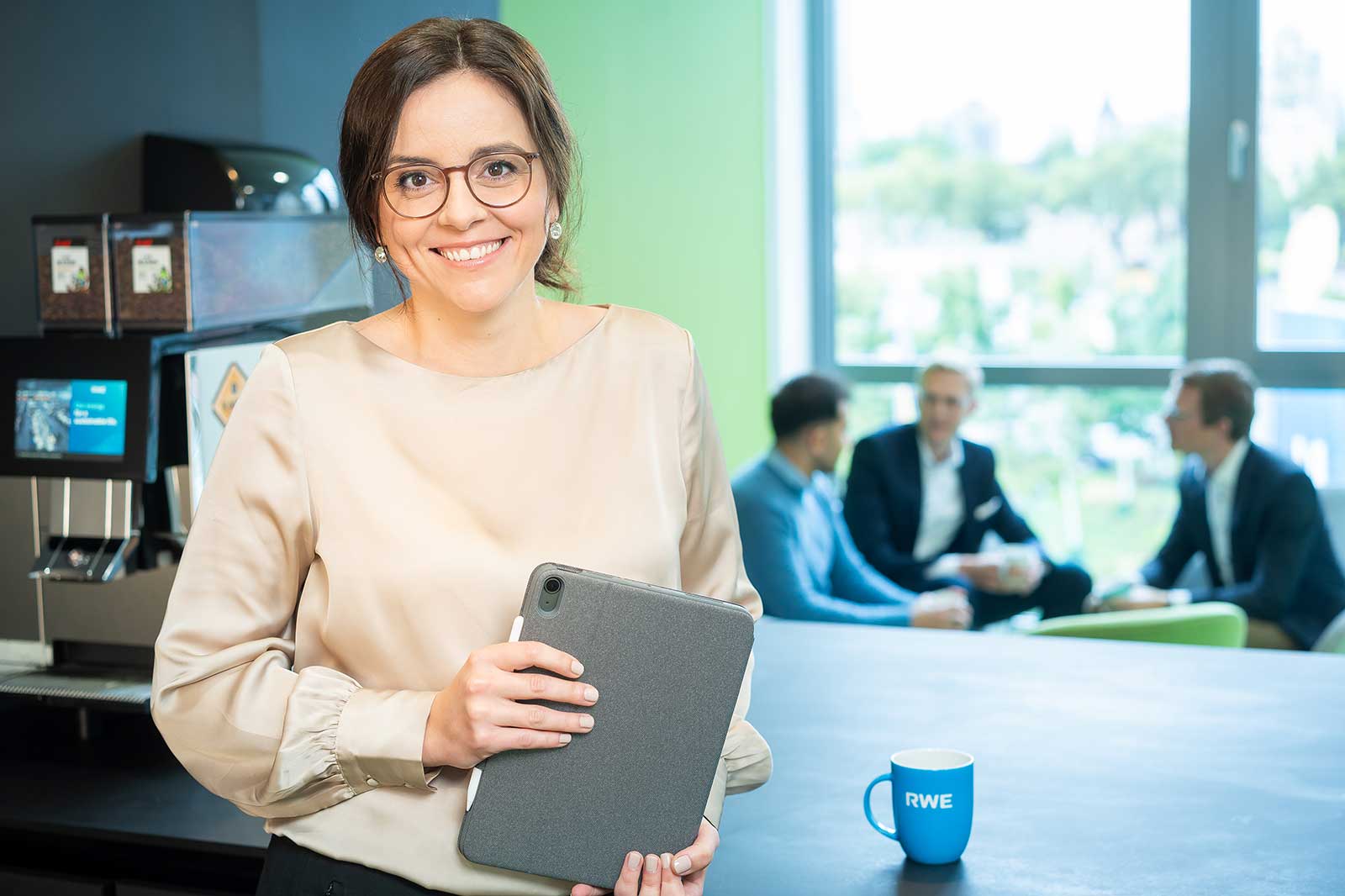 Eine Frau hält ein Tablet in einem modernen Büro, während eine Kaffeetasse mit dem Aufdruck RWE im Vordergrund steht und Kollegen im Hintergrund unterhalten.