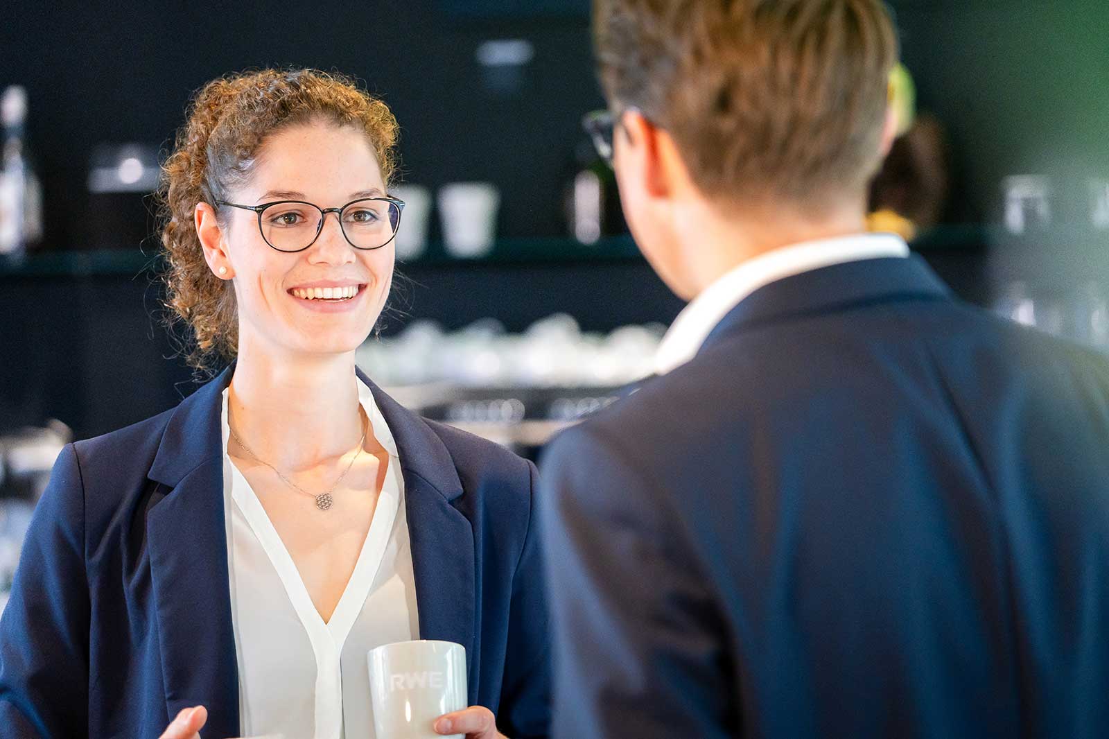 Eine Person in Anzug spricht mit einer anderen Person in einem Café mit Tassen im Hintergrund.