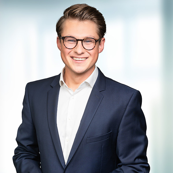 Ein junger Mann in einem dunkelblauen Anzug und einem weißen Hemd steht lächelnd vor einem hellen, unscharfen Hintergrund.
