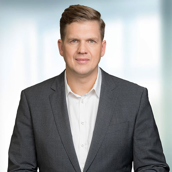 Ein Mann in einem Anzug und einem weißen Hemd steht mit neutralem Gesichtsausdruck vor einem sanft verschwommenen Hintergrund.