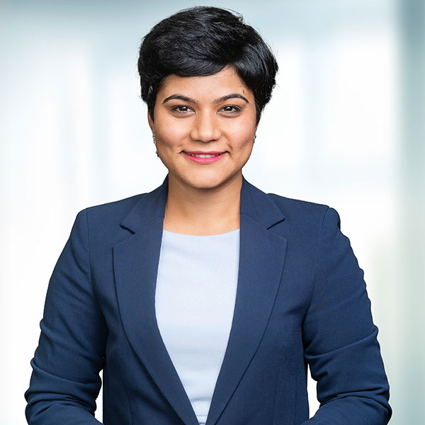Eine Person in einem dunklen Blazer und einem hellen Oberteil steht selbstbewusst mit verschränkten Armen vor einem verschwommenen Hintergrund.