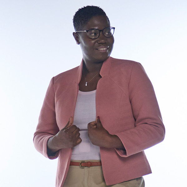 A person wears a light pink jacket over a white top. They pose with both hands on their waist.