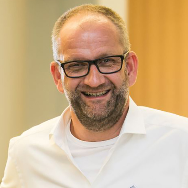 A man is wearing a white shirt and is depicted in a modern room with plain wooden walls in the background.