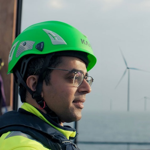 Ein Mann mit einem leuchtend grünen Helm steht am Wasser, während Windkraftanlagen im Hintergrund sichtbar sind.