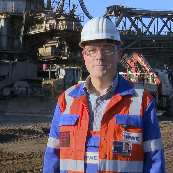 Ein Arbeiter in orange-blauem Sicherheitsanzug und Helm steht vor schweren Maschinen und Fahrzeugen auf einem industriellen Gelände.