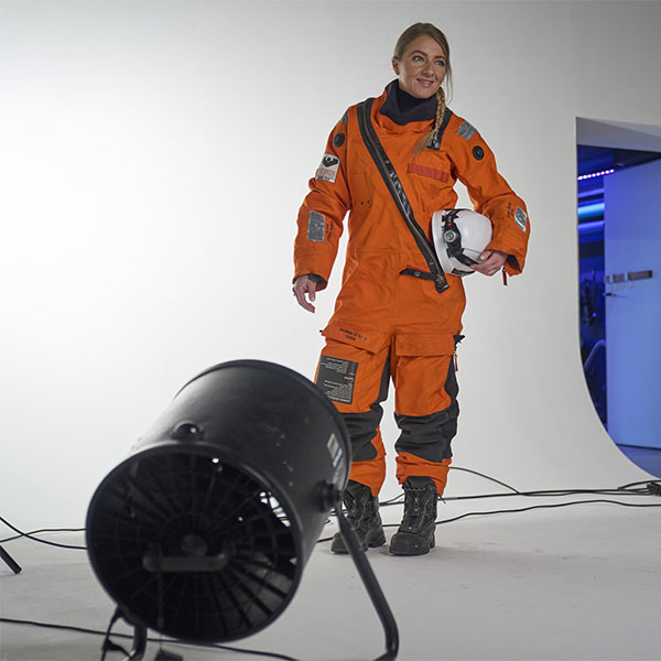 Eine Person in einem orangefarbenen Raumanzug hält einen Helm. Im Vordergrund befindet sich ein Ventilator.