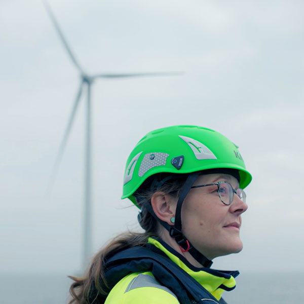 Eine Person mit einem grünen Helm und einer gelben Jacke steht vor einem Windrad an einem bewölkten Tag.
