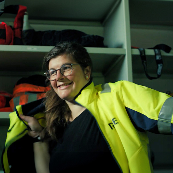 Eine Person trägt eine leuchtend gelbe Jacke, während ein Regal mit Rucksäcken im Hintergrund sichtbar ist.