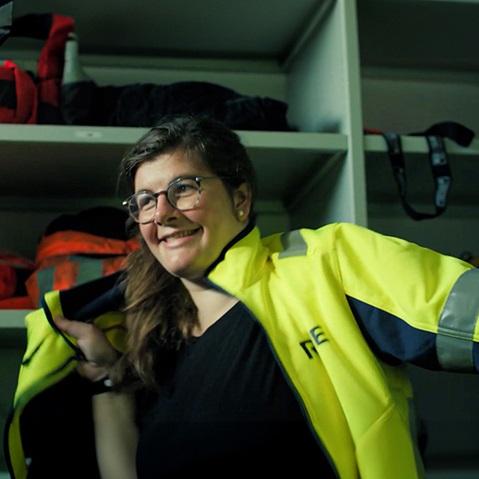 Eine Person trägt eine leuchtend gelbe Jacke, während ein Regal mit Rucksäcken im Hintergrund sichtbar ist.