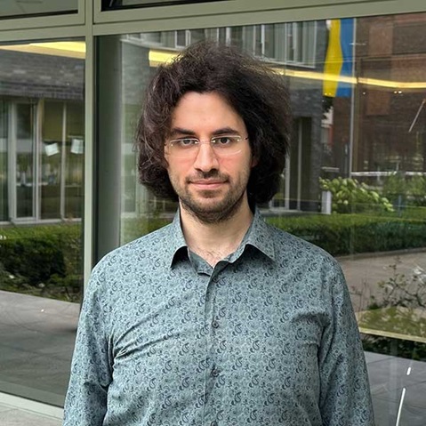 Ein Mann mit lockigem Haar trägt ein gemustertes Hemd und steht vor großen Fenstern in einem modernen Gebäude, Portrait von Dr. Farshid Balaneji.