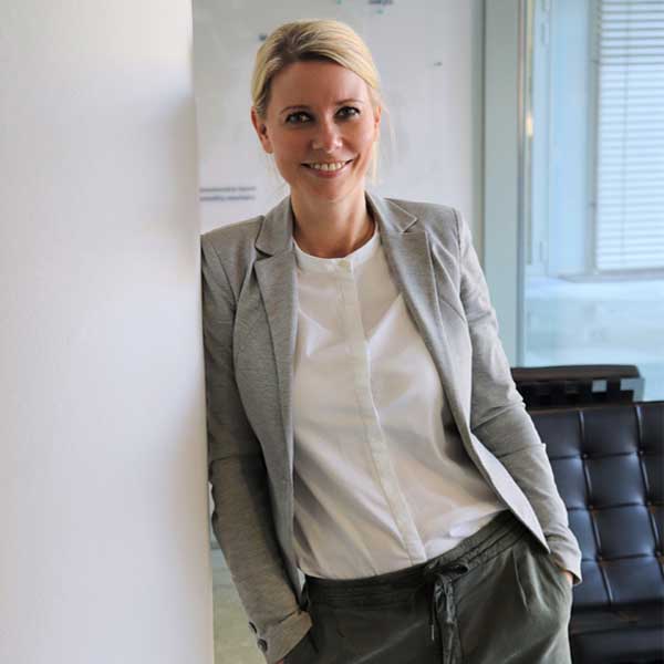 Eine Person in einem grauen Blazer und einer weißen Bluse lehnt an einer Wand in einem modernen Büro, Portrait von Madlen Krembel.