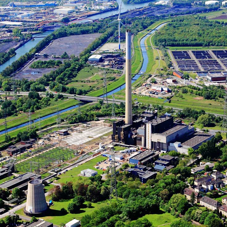 Luftaufnahme eines Industriegebiets mit einem Kraftwerk, Kühlturm und umliegendem Grün sowie Gewässern.