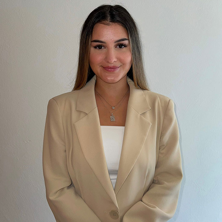 Eine Frau in einem beigen Blazer und einem weißen Oberteil steht vor einer hellen Wand und trägt Halsketten.