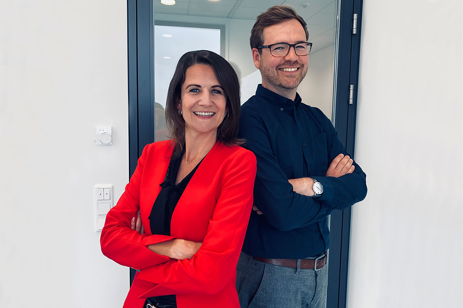 Ein Mann und eine Frau stehen mit verschränkten Armen in einem modernen Büro vor einer Glasscheibe.