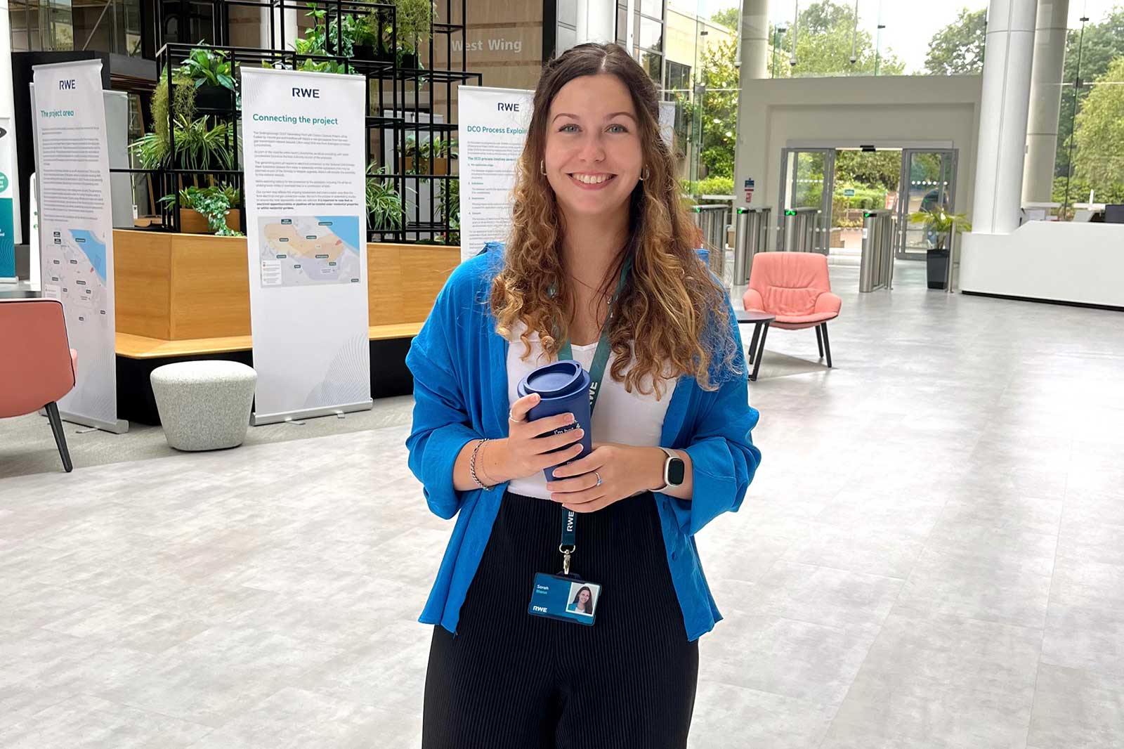 A person stands in a modern space with plants and information stands. They hold a cup and wear a blue jacket.