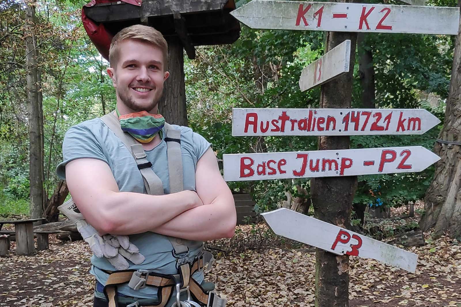 A man stands with crossed arms in front of a signpost in the woods, pointing to various adventure activities.