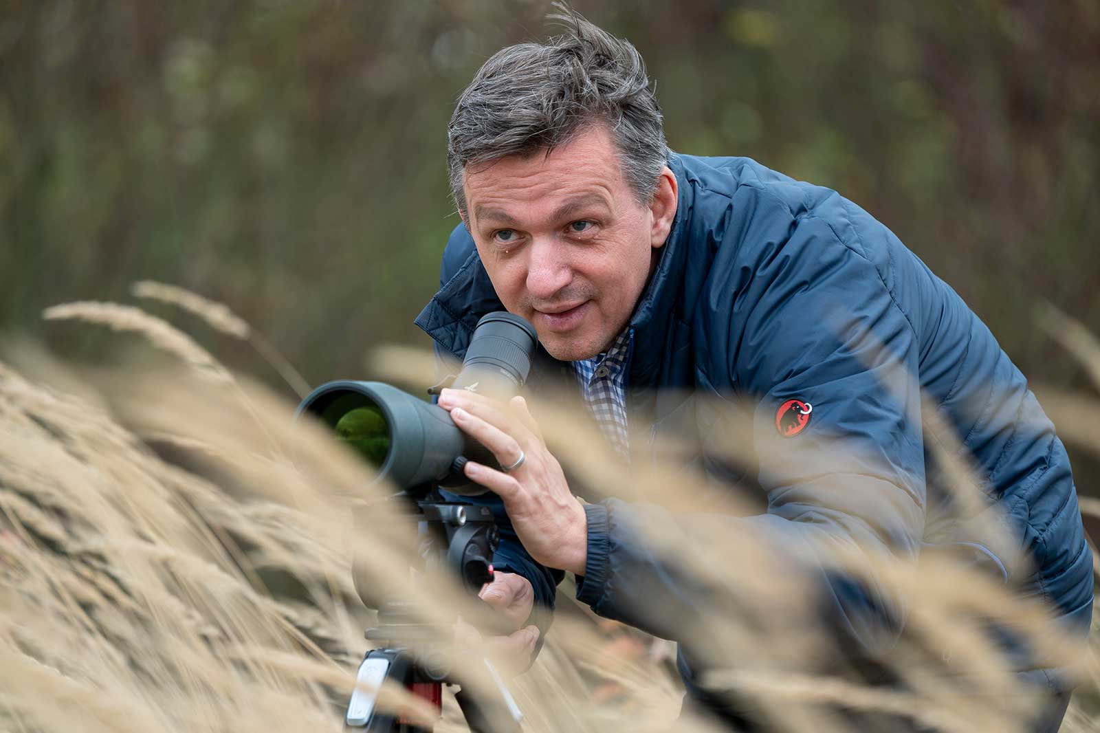 Ein Mann mit einem Fernrohr sitzt im hohen Gras und beobachtet die Umgebung aufmerksam.