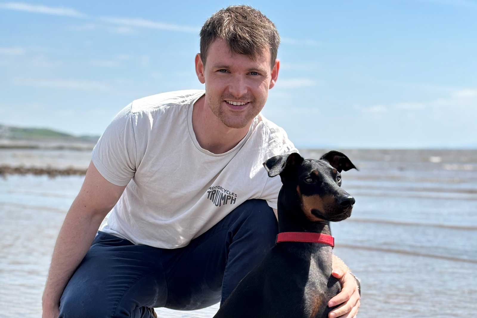 Ein Mann kniet am Strand neben einem schwarzen Hund. Der Himmel ist blau und die Wellen sind sanft.