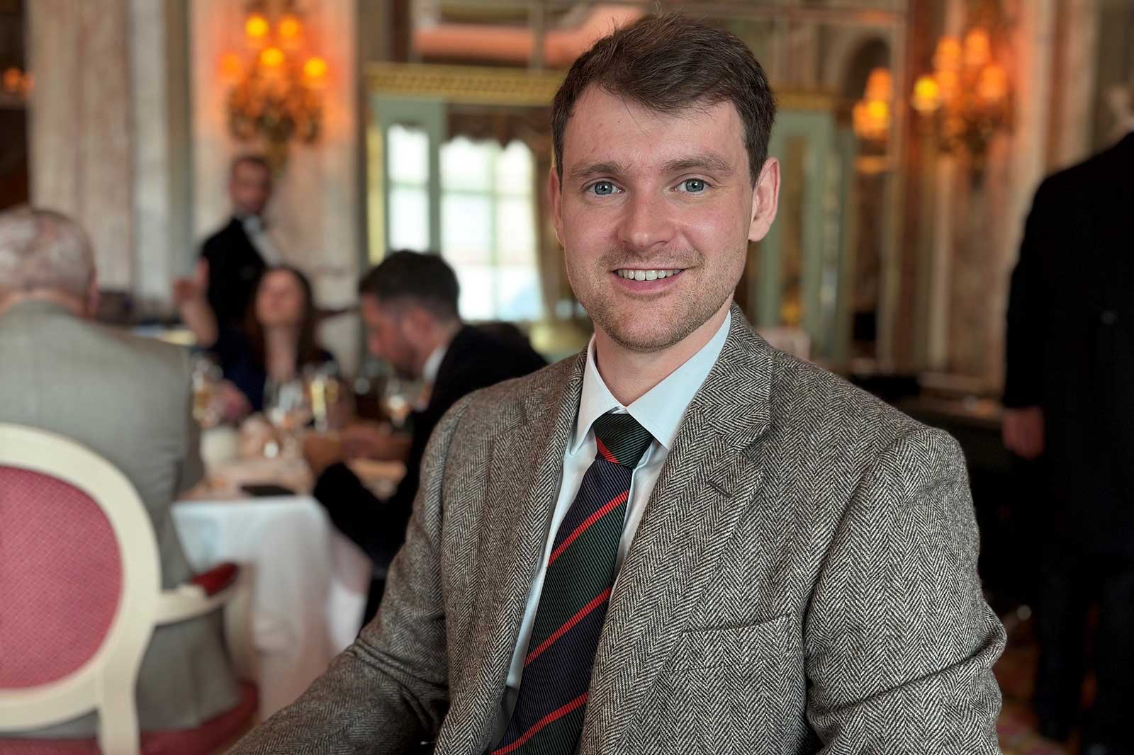 Ein Mann in einem eleganten Restaurant, in einem grauen Tweed-Anzug mit einem gestreiften Krawatte, sitzt an einem Tisch.