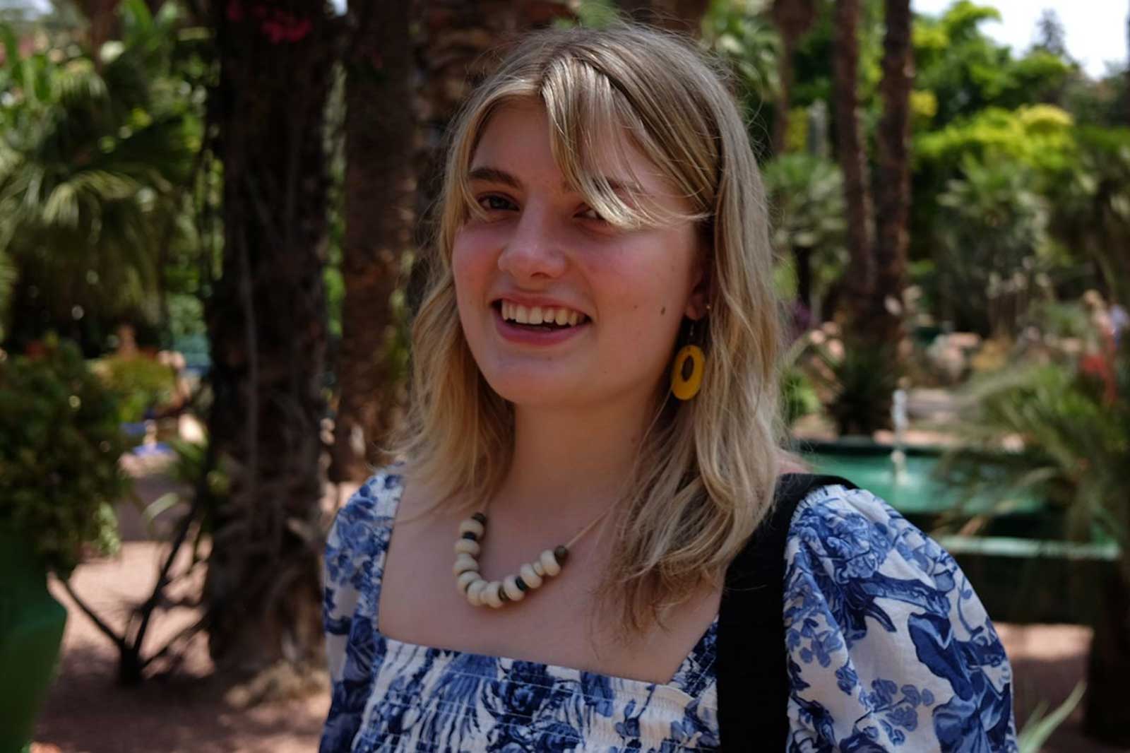 A woman with blonde hair is wearing a patterned blouse and a necklace. The background features palm trees and greenery.