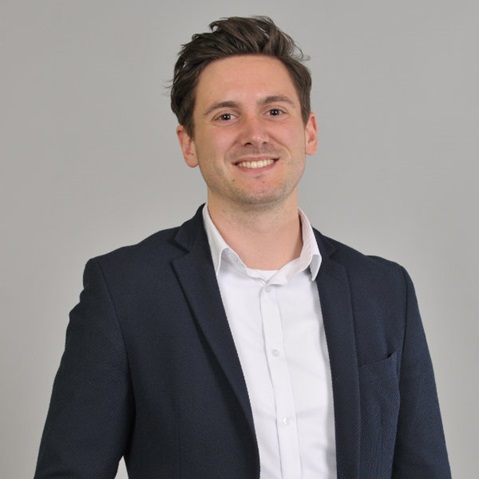 Ein Mann in einem schwarzen Anzug mit einem weißen Hemd steht vor einem grauen Hintergrund.