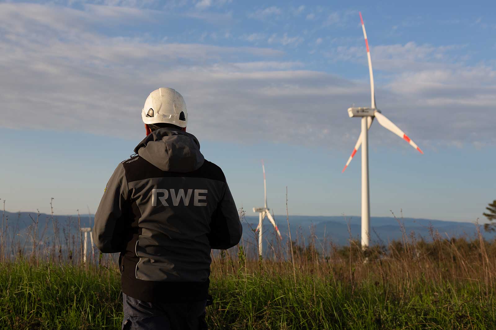 Ein Mitarbeiter mit Schutzhelm und RWE-Jacke steht auf einem grasigen Hügel und blickt auf Windräder vor blauem Himmel und fernen Hügeln.