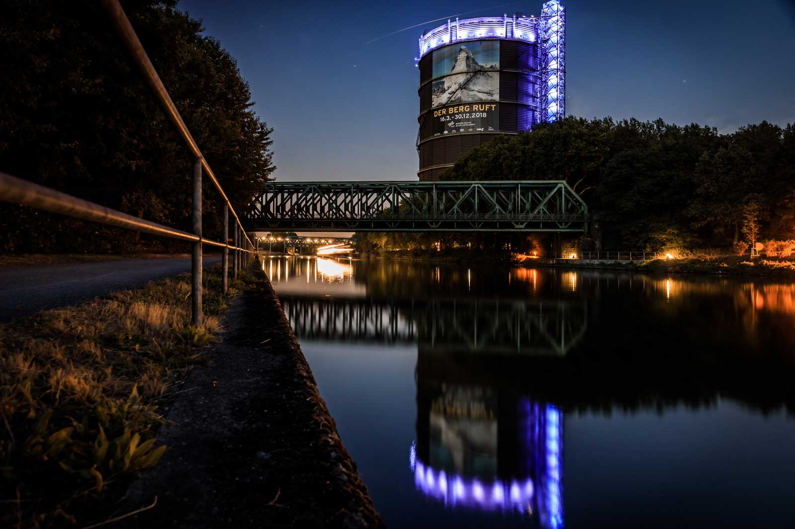 Eine beleuchtete Industrieanlage am Wasser bei Nacht, neben einer grünen Brücke und einem asphaltierten Weg.
