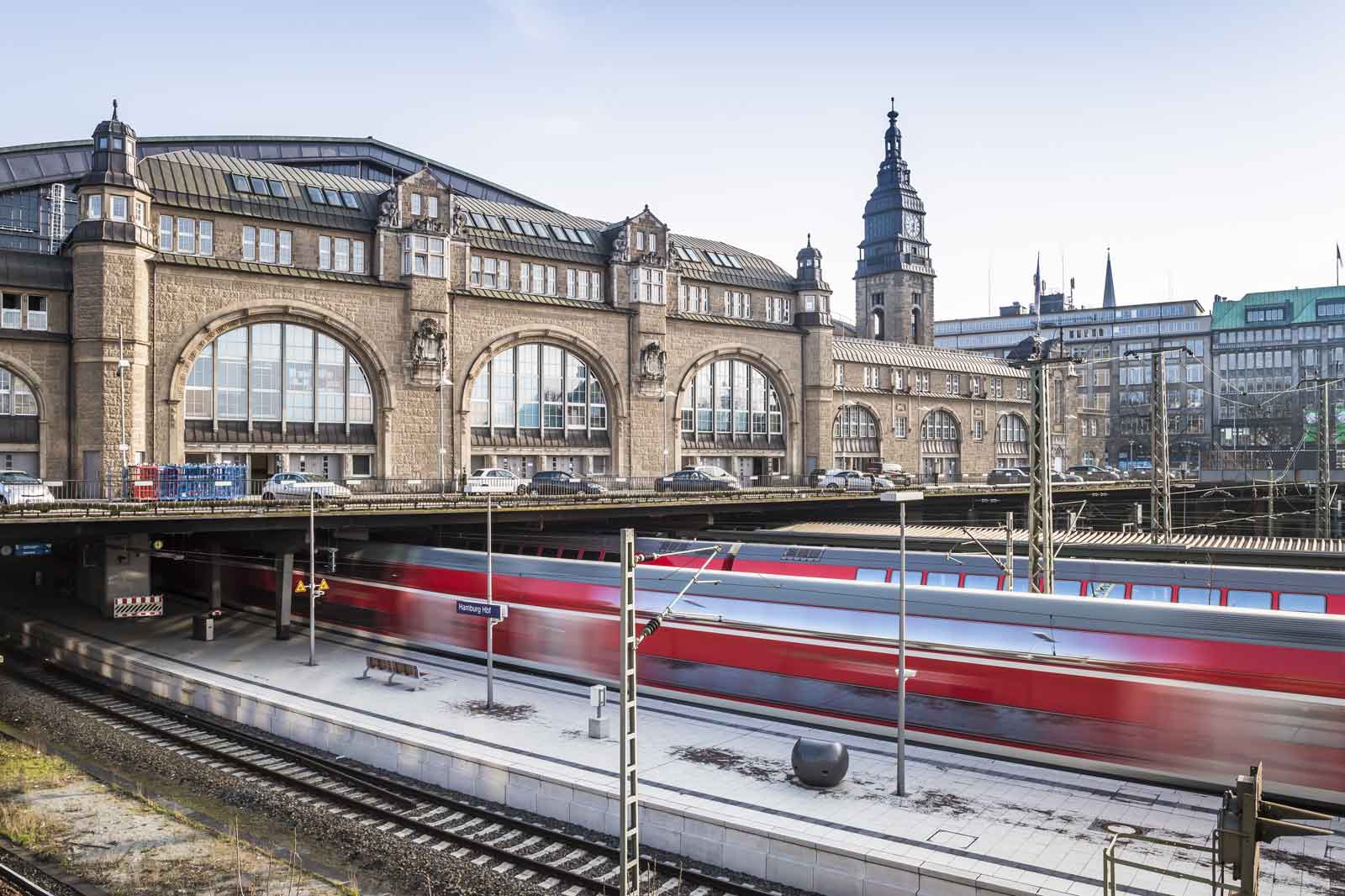 Ein historischer Bahnhof mit beeindruckender Architektur, während ein roter Zug vorbeifährt.