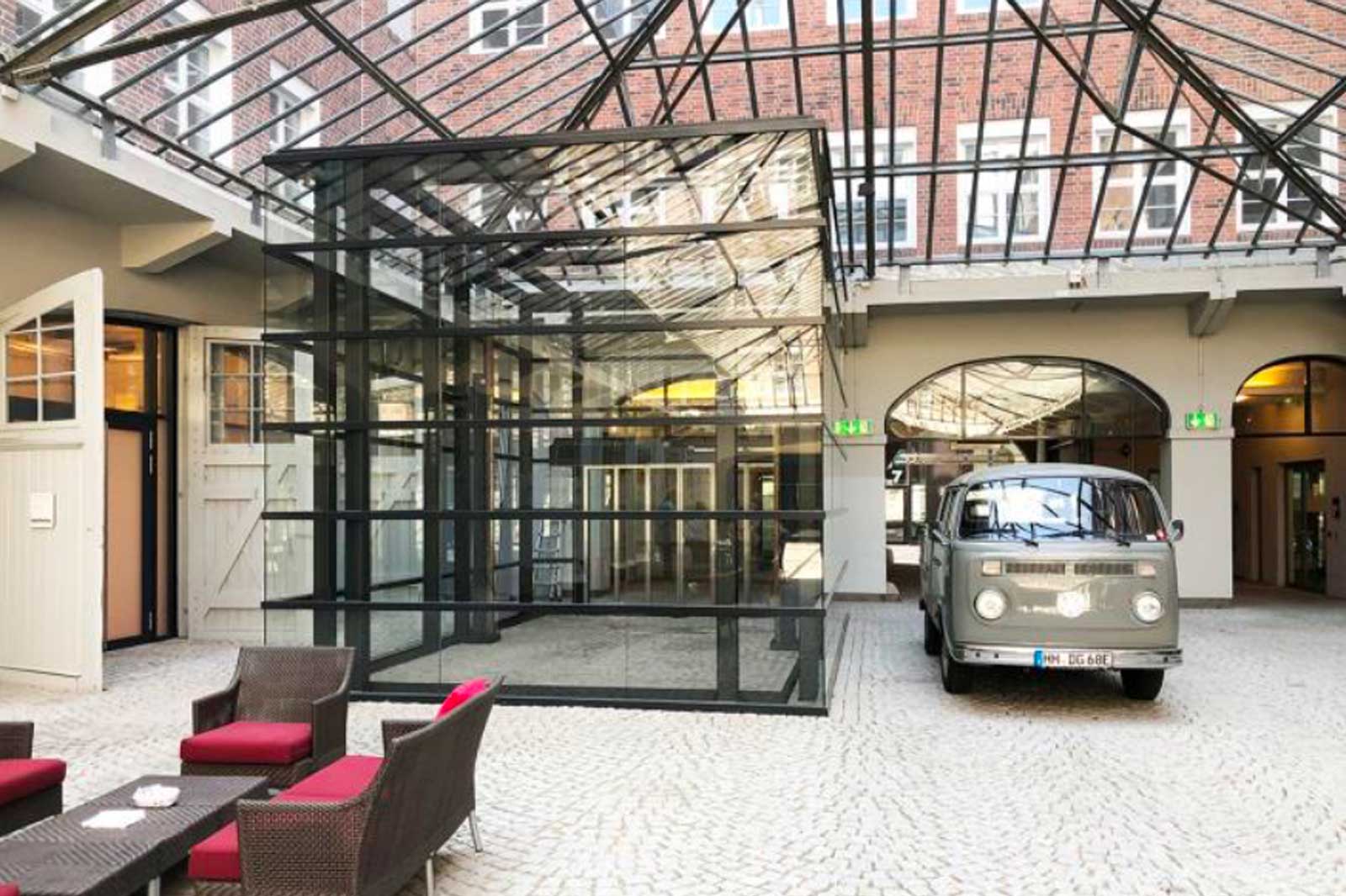Ein modernes, gläsernes Gebäude im Innenhof mit einem alten Lieferwagen und einem Sitzbereich mit roten Kissen.
