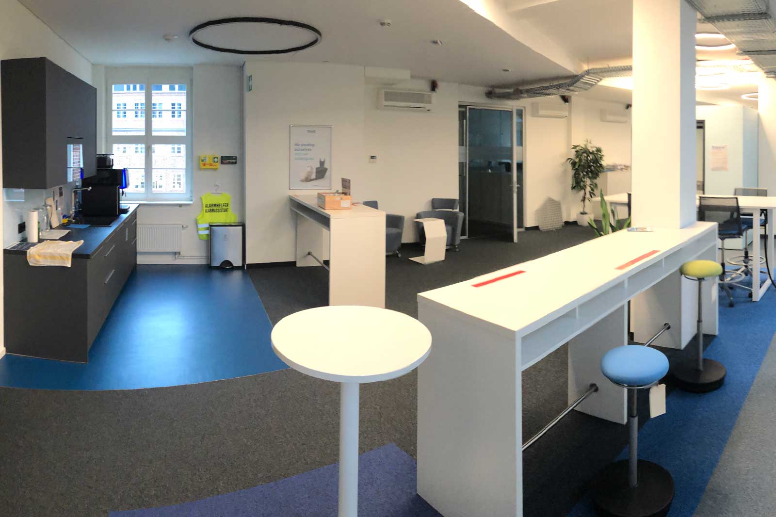 Das Bild zeigt ein modernes Büro mit einer Kaffeeküche, Tischen, Stühlen und Pflanzen im Hintergrund.