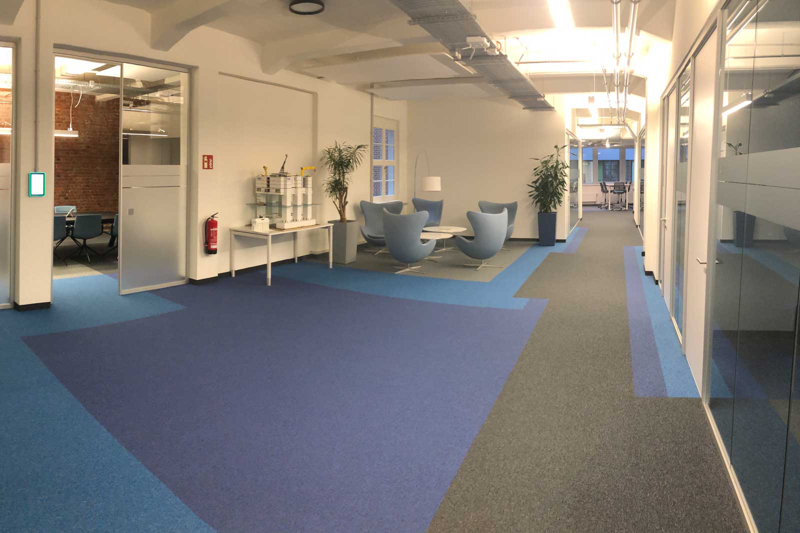 Ein modernes Büro mit blauen Teppichen, gemütlichen Sesseln und Pflanzen. Ein Besprechungsraum ist sichtbar.