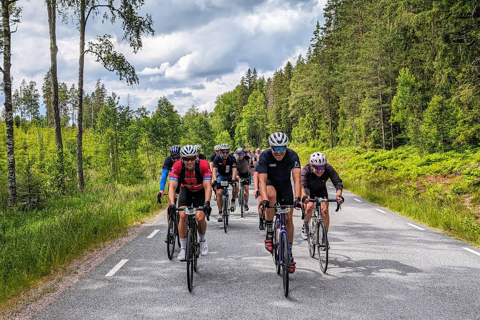 Eine Gruppe von Radfahrern fährt auf einer Landstraße, umgeben von grünen Bäumen und einem bewölkten Himmel.